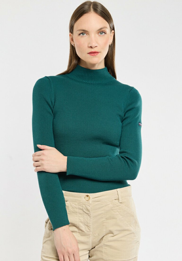 Maglione a collo alto in teal a coste con maniche lunghe. Texture morbida e design aderente. Indossato con pantaloni beige chiaro. Logo significativo sulla manica.