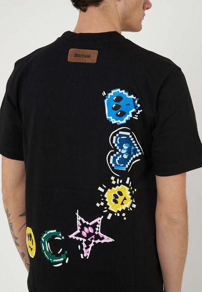 Man wearing black T-shirt with colorful pixel-style graphics including blue skull, blue heart, yellow sun, pink star, and yellow smiley on back right side.