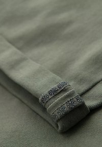 Un tissu vert avec une surface texturée présente un ourlet replié. Le poignet est orné d'un motif de surpiqûre décorative bleue.