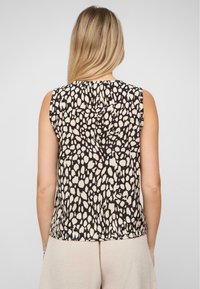 Femme avec de longs cheveux blonds porte un haut sans manches noir et beige à pois et un pantalon beige clair devant un fond uni.