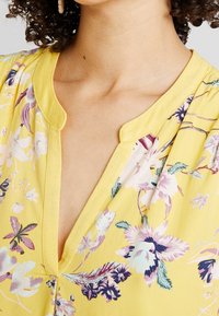 Gelbe Bluse mit Blumenmuster, V-Ausschnitt, leichtem Stoff und einer Mischung aus lila, blauen und grünen Blumenmustern.