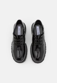 Paire de chaussures à lacets en cuir verni noir avec semelles épaisses à crampons et étiquette de marque à l'intérieur sur fond blanc.