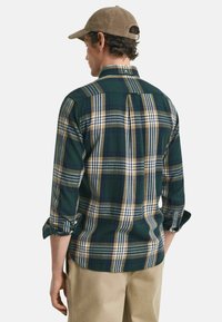 Groen en marineblauw geruit overhemd met gele en witte strepen, lange mouwen opgerold, achterste naad en een afgeronde onderkant. Draagt met khaki broek.