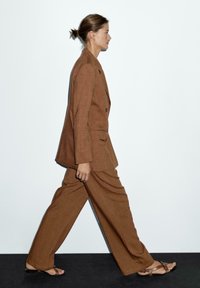 Bruin linnen pak met wijde pijpen en een getailleerde blazer, gecombineerd met platte sandalen. Minimalistisch ontwerp tegen een effen witte achtergrond.