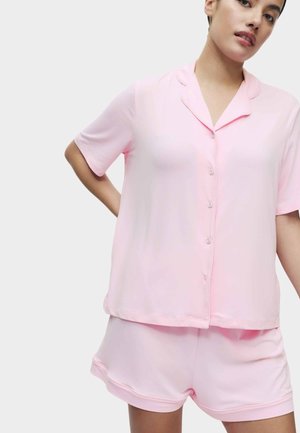 Kobieta ubrana w jasnoróżową, krótki rękaw, zapinaną na guziki bluzkę i dopasowane krótkie spodenki, stojąca na jednolitym białym tle.