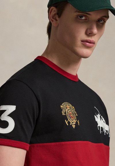 Νέος άνδρας που φοράει μαύρο και κόκκινο πόλο με κεντημένα λογότυπα και πράσινη καπέλο μπέιζμπολ, κοιτάζοντας την κάμερα.