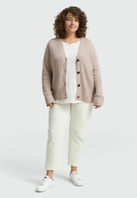 Cardigan en tricot beige clair avec texture côtelée, fermeture à boutons et manches larges, porté sur un haut blanc et assorti avec un pantalon blanc et des baskets.