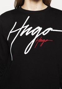 Černá mikina s kulatým výstřihem, na které je velký bílý kurzívní nápis "Hugo" a pod ním menší červený kurzívní nápis "Hugo" vyšité na hrudi.