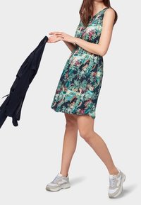 Ärmlös klänning med ett tropiskt mönster som innehåller gröna, rosa och blå mönster. Mörkblå tyg hålls i ena handen; grå och vita sneakers på fötterna.