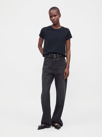 Femme aux cheveux courts portant un t-shirt noir, un jean foncé taille haute avec une ceinture à motif, et des chaussures noires à bout pointu.