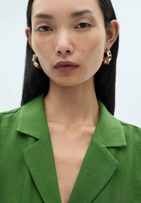 Zelený lněný blazer s klopovým límcem, s texturovaným povrchovým zpracováním. Model nosí zlaté kruhové náušnice, má hladkou pleť a rovné černé vlasy.