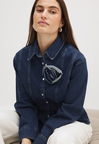 Dark denim shirt with a structured collar, button-up front, and a frayed denim rose accent on the chest. Long sleeves with a relaxed fit.