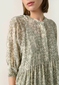 Blusa ligera y transparente en un patrón abstracto de verde apagado y beige, con cuello redondo y mangas tres cuartos con fruncidos.