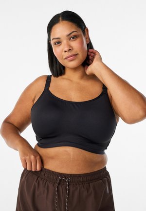 Keskmise nahatooni ja sirgete mustade juustega naine kannab musta spordirinnahoidjat ja pruune nööriga pükse, vaatab kaamerasse käsi kaelal.