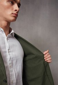 Zielona kurtka o strukturalnym fasonie, połączona z białą lnianą koszulą, prezentująca boczną kieszeń i detale z guzikami. Teksturowane wykończenie tkaniny.