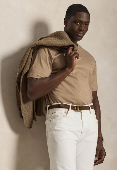 Bezsínű pólót és fehér nadrágot viselő férfi, aki egy barna halszálka mintás zakót tart az egyik vállán, semleges háttér előtt.