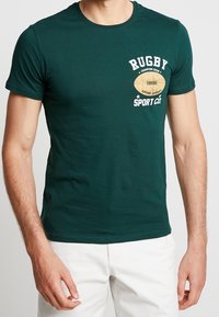 Camiseta de algodón verde con escote redondo. Presenta un gráfico de balón de rugby y el texto "RUGBY" en blanco, sobre un óvalo color crema con "SPORT CO."