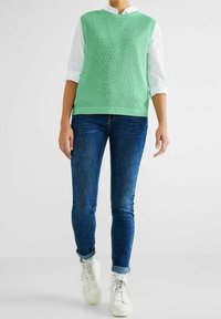 Light green knitted sleeveless top over a white collared shirt, paired with blue skinny jeans and white sneakers. Simple, casual outfit.
