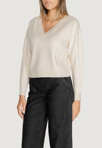 Maglione crema a V con maniche lunghe e orlo a coste, abbinato a pantaloni grigi scuri. Tessuto liscio, vestibilità rilassata.