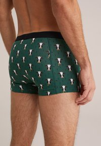 Man som bär mörkgröna boxerkalsonger mönstrade med små svartvita kofröner, stående med ryggen och låret synligt.