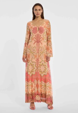 Vrouw die een lange, perzikkleurige jurk met bloemenprint draagt, met roze, groene en oranje patronen en roze open schoenen, staand tegen een witte achtergrond.