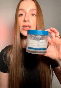 Donna con capelli lunghi che tiene in mano un barattolo di Christophe Robin Cleansing Purifying Scrub per cuoio capelluto sensibile o grasso, parlando alla telecamera.