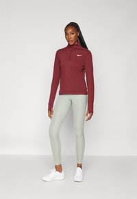 Burgundi színű hosszú ujjú felső, negyed zippel és Nike logóval, világoszöld magas derekú leggings-szel és fehér sportcipővel párosítva.
