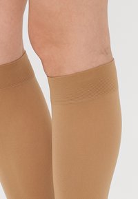 Ciorapi de compresie până la genunchi, de culoare bej deschis, confecționați dintr-un material neted și elastic, cu vârfuri ribbate. Potriviți pentru purtare zilnică.