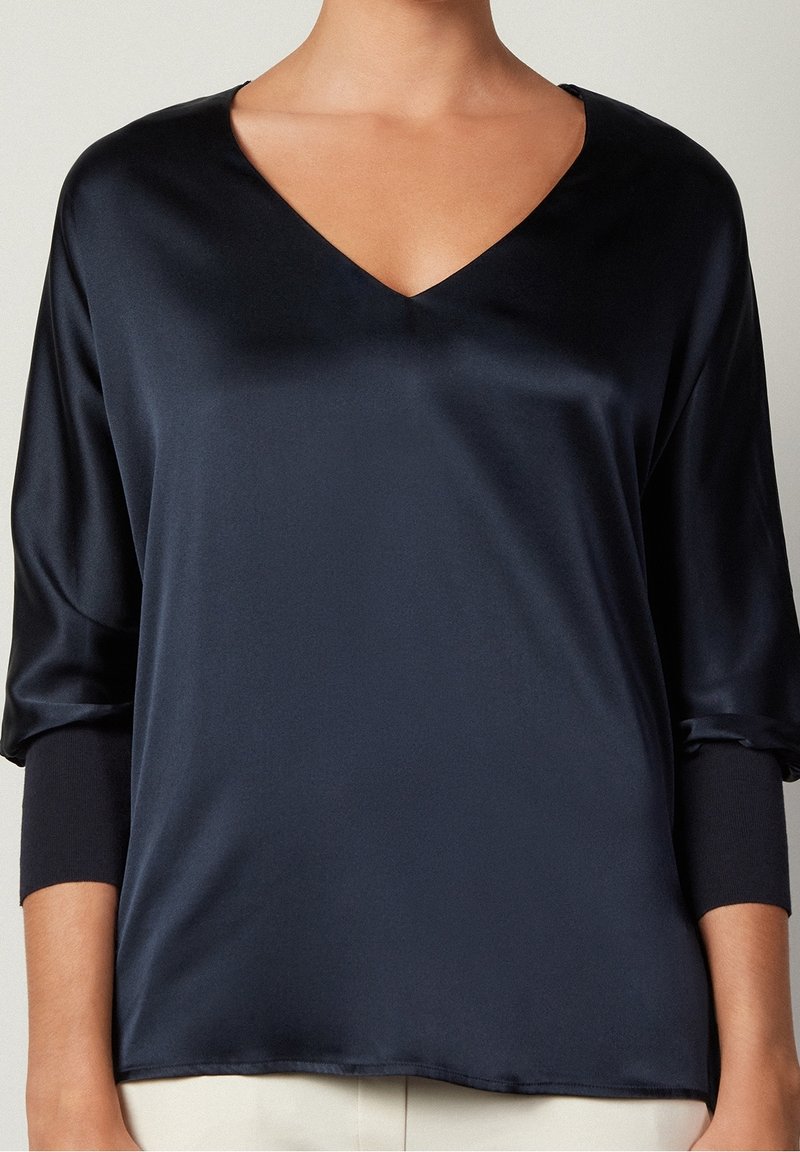 Blouse en satin bleu marine avec un col en V, des manches trois-quarts et une texture lisse. Le tissu tombe doucement et semble léger.