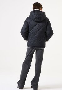 Chaqueta acolchada negra con capucha y detalle de piel sintética, con un patrón curvado. Combinada con pantalones de pernera ancha grises.