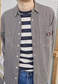 Camicia a righe bianche e nere con due tasche sul petto, indossata sopra una maglietta a girocollo a righe blu e bianche, abbinata a jeans blu.