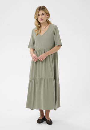 Kobieta ubrana w luźną, jasnooliwkową sukienkę midi z falbanami i krótkimi rękawami, nosząca czarne plecione baleriny, stoi na tle jednolitym.