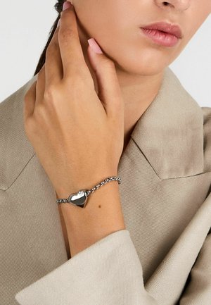 Bracciale a cuore in argento con design a catena. Il pendente a cuore presenta un piccolo accento di diamante, indossato su una mano contro un tessuto neutro.