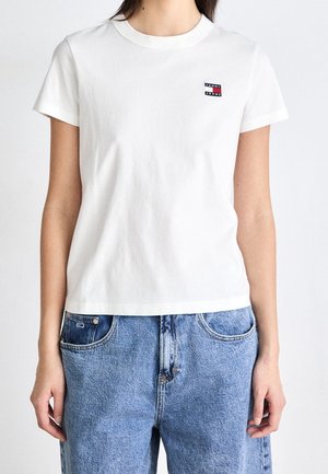 Kobieta nosząca gładką białą koszulkę z krótkim rękawem z małym prostokątnym logo Tommy Jeans na piersi, zestawioną z jasnoniebieskimi jeansami.