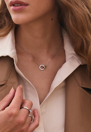Sölvhalsband med cirkulärt hänge, prytt med små klara stenar. Modellen bär en beige kappa över en ljus blus, med ringar på sina fingrar.
