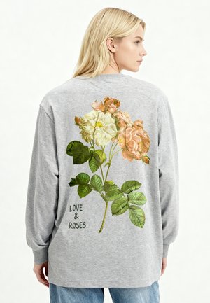 Femme blonde portant un t-shirt gris à manches longues avec un grand imprimé floral et le texte "LOVE & ROSES" dans le dos, vue de profil.