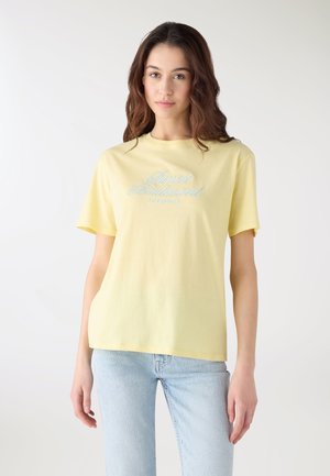 Giovane donna con capelli castani ondulati, indossa una maglietta a maniche corte giallo chiaro con testo azzurro chiaro e jeans azzurri chiari, in piedi davanti a uno sfondo bianco.