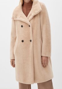 Beige dubbelknäppt kappa av mjukt, texturerat tyg med bred krage, svarta knappar och sidofickor, bärs över en brun polotröja.