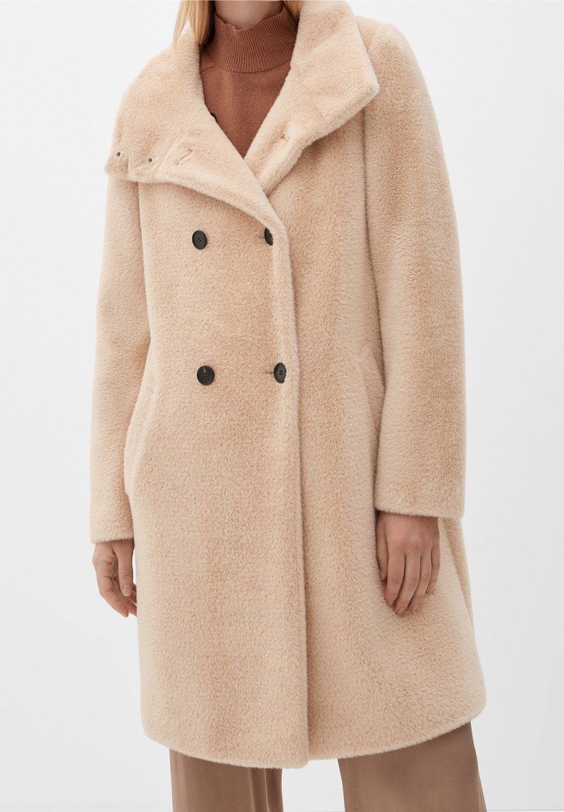 Beige dubbelknäppt kappa av mjukt, texturerat tyg med bred krage, svarta knappar och sidofickor, bärs över en brun polotröja.