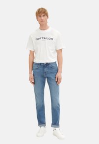 Tricou alb din bumbac cu text „TOM TAILOR” în nuanță de bleumarin, asortat cu blugi din denim de un albastru deschis și adidași albi. Croi simplu, casual.