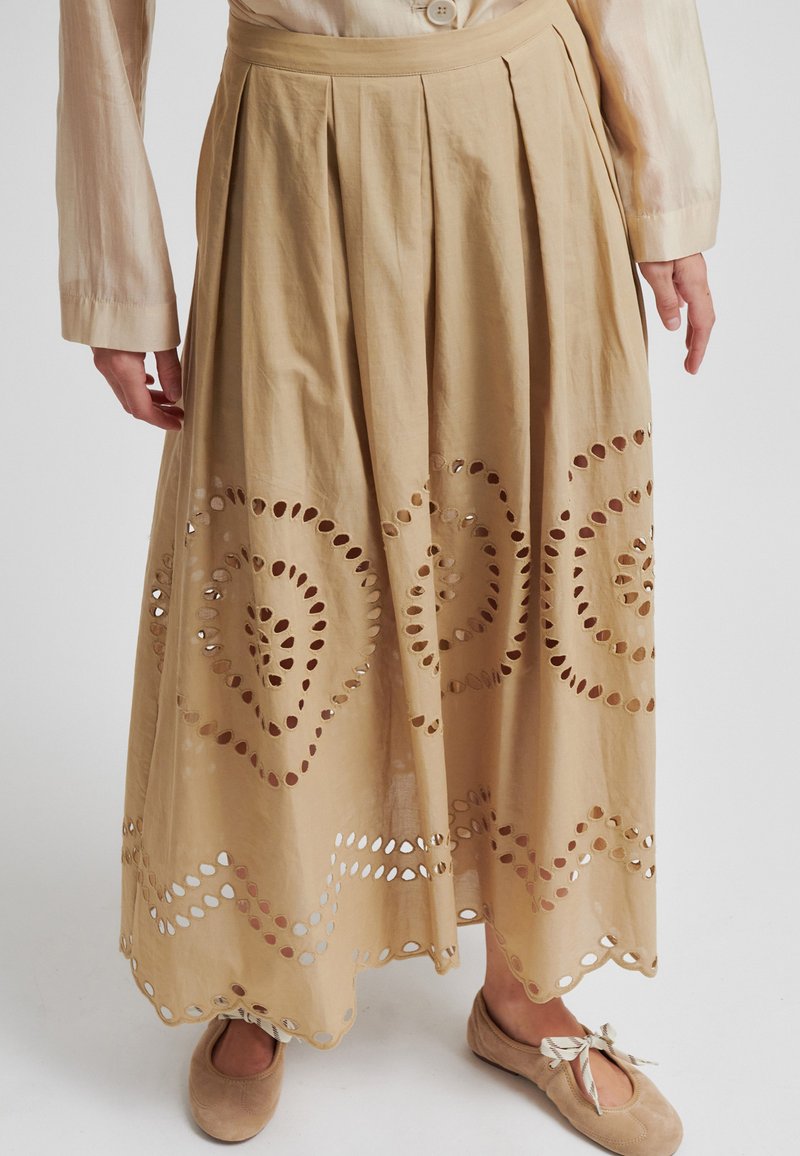Jupe midi plissée beige avec des motifs circulaires découpés complexes, portée avec un chemisier assorti et des chaussures plates beiges à détails nœud.