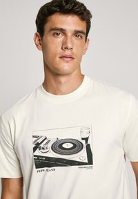 Krēms krāsas T krekls ar melnu grafiku, kurā attēlota vinila grozītāja un glāzes, ar tekstu "PEPE JEANS" un "PORTOBELLO RD" biezā, vienkāršā fontā.