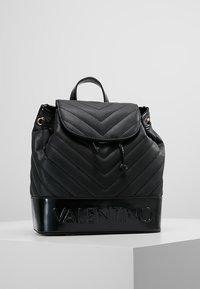 Musta nahkareppu, jossa on chevron-neliskulmainen kuvio, nauhakiristys ja logo yksityiskohtana pohjassa. Varustettu yläkahvalla ja säädettävillä olkahihnoilla.