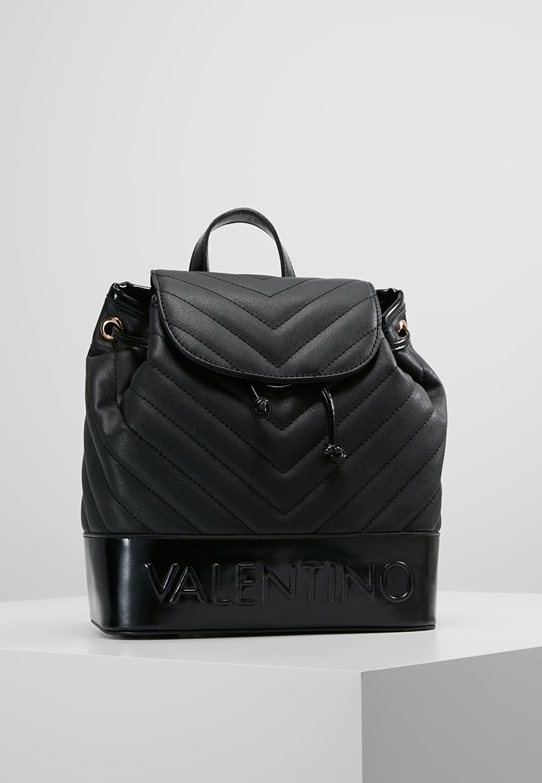 Musta nahkareppu, jossa on chevron-neliskulmainen kuvio, nauhakiristys ja logo yksityiskohtana pohjassa. Varustettu yläkahvalla ja säädettävillä olkahihnoilla.