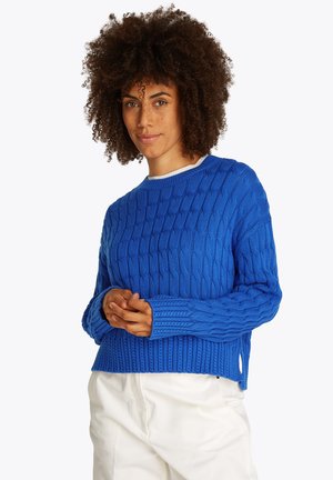 Frau mit lockigem Haar, die einen leuchtend blauen, grob gestrickten Pullover und weiße Hosen trägt, steht mit sanft gefalteten Händen.