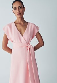 Rožinė, kimono stiliaus suknelė su trumpomis rankovėmis ir giliu V iškirpimu. Suknelė turi suvarstomą juosmenį ir glotnią, plaukiančią medžiagą su subtiliu blizgesiu.