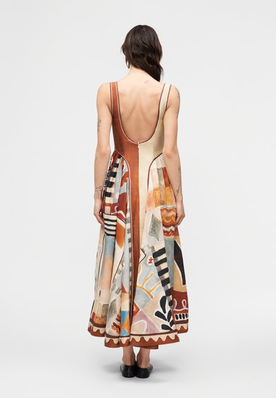 Γυναίκα με σκοτεινά μαλλιά φοράει αμάνικο φόρεμα με πολύχρωμο αφηρημένο μοτίβο και χαμηλή πλάτη, καθώς και μαύρα επίπεδα παπούτσια, στέκεται μπροστά σε απλό φόντο.