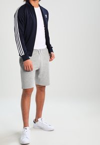 Giacca atletica blu navy con strisce bianche, t-shirt bianca, pantaloni corti grigi e scarpe da ginnastica bianche, caratterizzata da un design sportivo e una vestibilità rilassata.