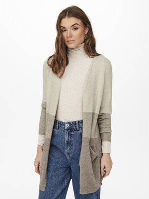 Beiger Strickcardigan mit offenem Vorderteil und zwei Taschen, mit einem Farbverlauf von hellgrau zu dunkelgrau, getragen über einem enganliegenden cremefarbenen Rollkragenpullover.