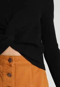 Svart ribbad långärmad topp med knutdesign fram, parat med en orange kordflossholkskjorta med knappstängningar.